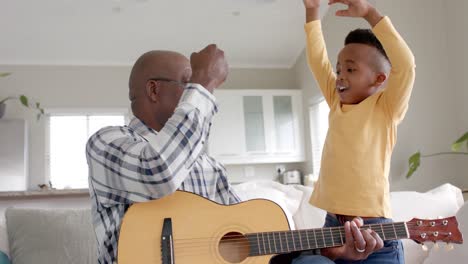 Happy-african-american-grandfather-and-grandson-playing-guitar,-fist-bumping-at-home,-slow-motion
