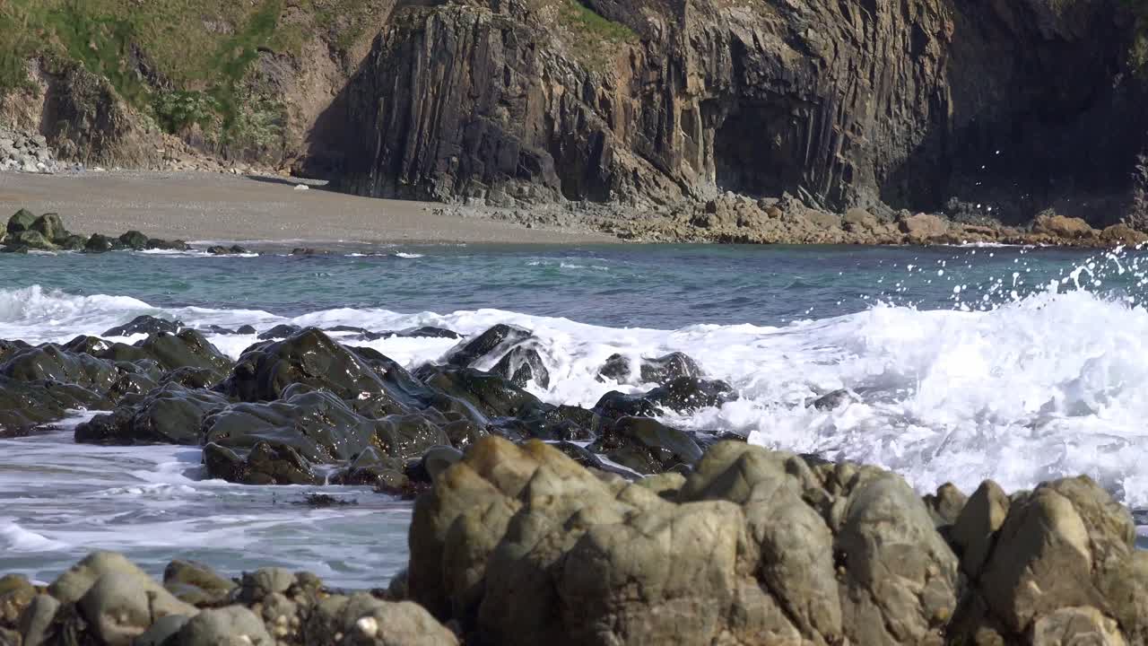 Onde che si infrangono su una spiaggia della Copper Coast di Waterford con rocce di tutti i colori e texture, una costa deserta in una calda mattina d'estate.