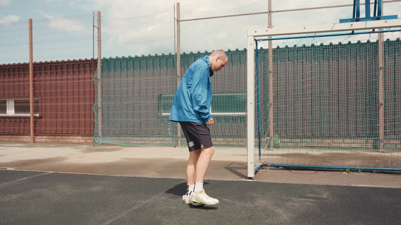Bílý fotbalista na mokrém městském hřišti, modrá bunda se leskne deštěm, odráží míč u brankové tyče poblíž zrezivělého plotu a betonové plochy. Samostatná tréninková sekvence ukazuje hráče, jak se rozcvičuje a kontroluje síť.