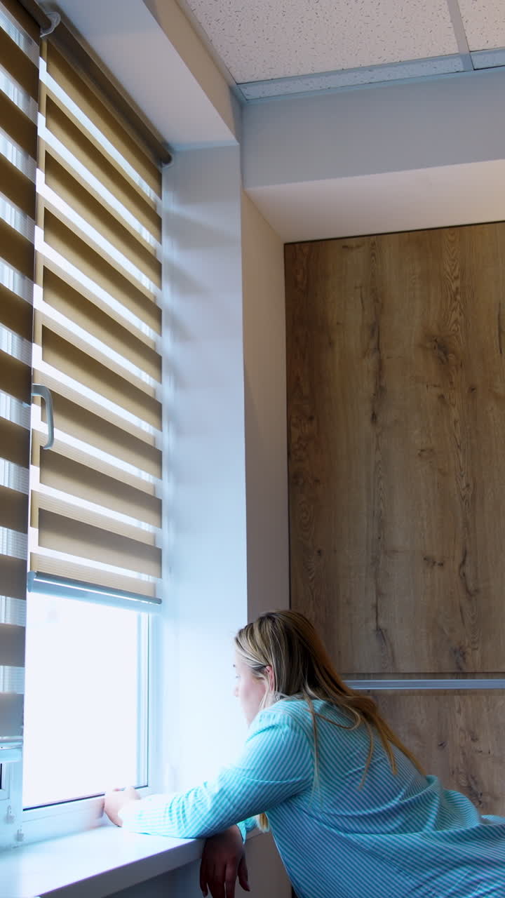 Caucasian woman stands leaned on the window-sill looking into window. Future mom in the maternity hospital ward. Vertical video