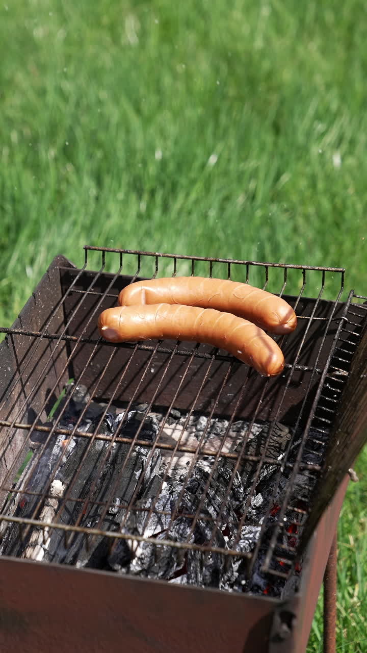 Man puts sausages into a grill for frying. Cooking hot dog sausages on fire. Barbecue in nature. Brazier on green grass background. Vertical video