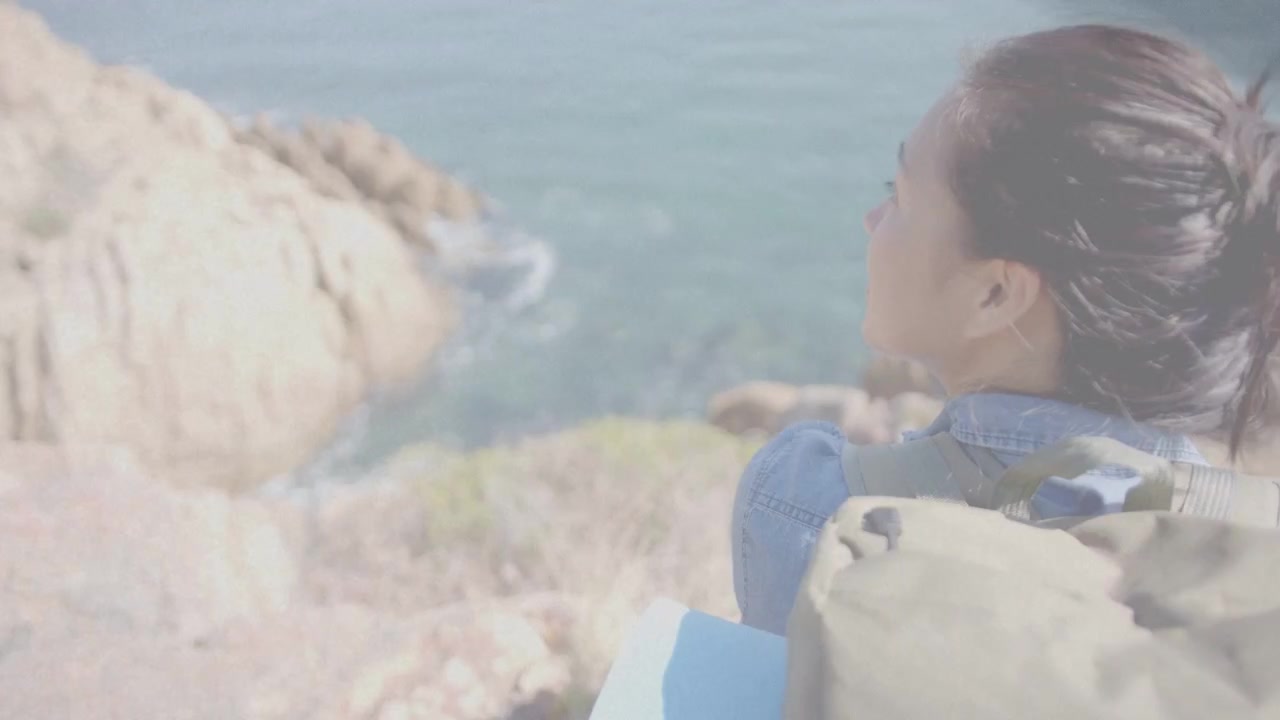 崖で立ち止まり海を眺めるハイカー。カメラが安定して海岸線が映し出され、タン色のリュックと青いマットを身につけている。旅。