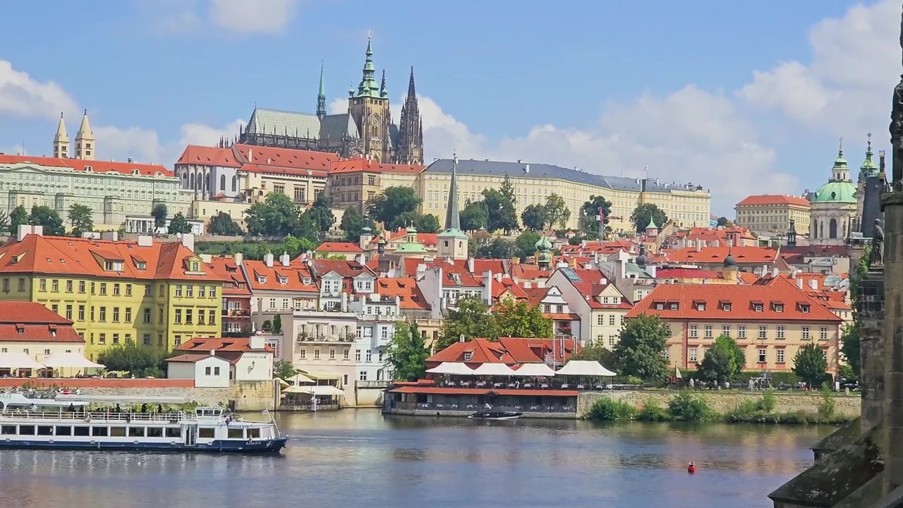 Paesaggio urbano di Praga dall'altra parte del fiume Moldava, che mostra la collina del castello, la Cattedrale di San Vito, tetti di tegole arancioni, ristoranti sul lungofiume, un battello turistico e un luminoso scenario di viaggio europeo.