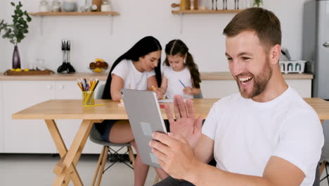 hombre guapo feliz que tiene una videollamada usando una tableta digital en casa mientras en el fondo su esposa y su pequeña hija se sientan a la mesa y dibujan juntas