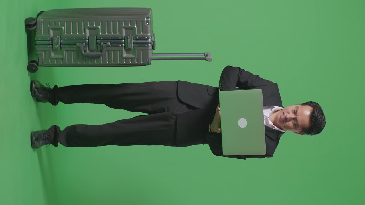 cuerpo lleno de viajero hombre de negocios asiático con equipaje usando una computadora portátil mientras estaba de pie en el estudio de fondo de la pantalla verde