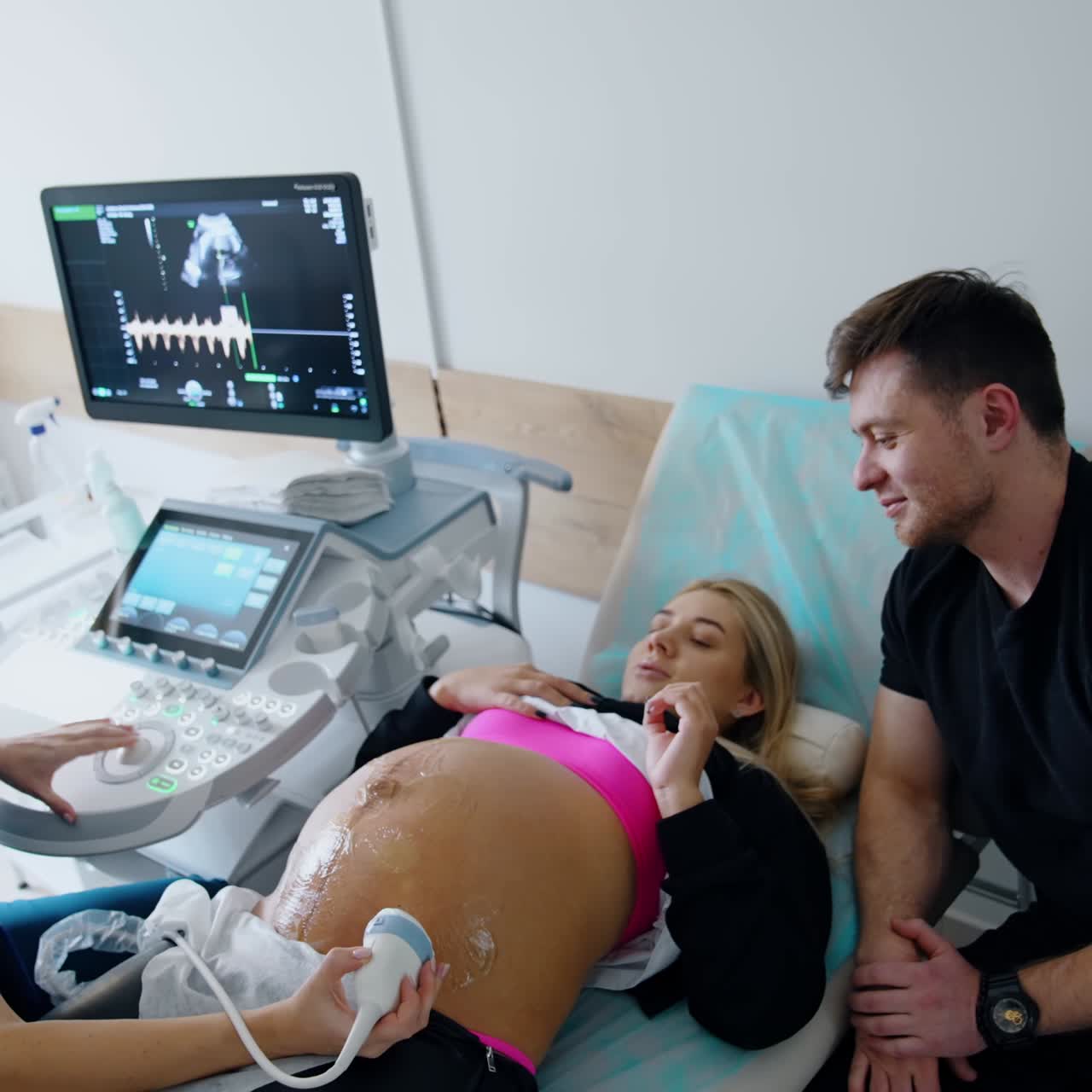 Donna incinta parla con il medico durante un esame ecografico. Un uomo sorridente è seduto accanto ad ascoltare. Vista dall'alto