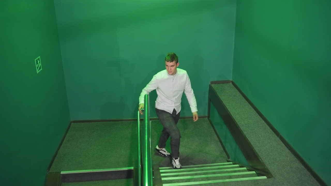 hombre con camisa y pantalones subiendo las escaleras, sosteniendo el riel con una expresión seria bajo la iluminación verde. las sombras proyectadas en las paredes crean una atmósfera de mal humor en el interior moderno con barandillas metálicas elegantes