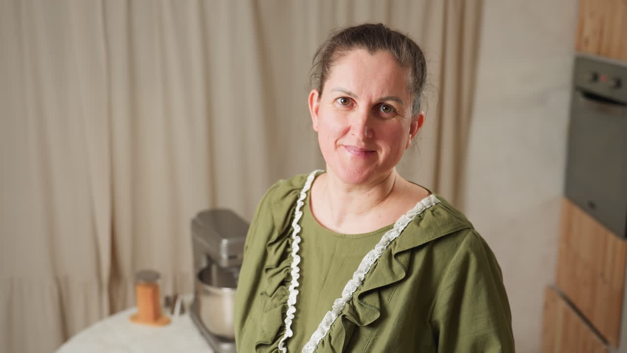 Portrait view of woman in green ruffled gown looking calmly at camera with soft expression, standing in cozy kitchen with electric mixer on table behind, light curtain backdrop
