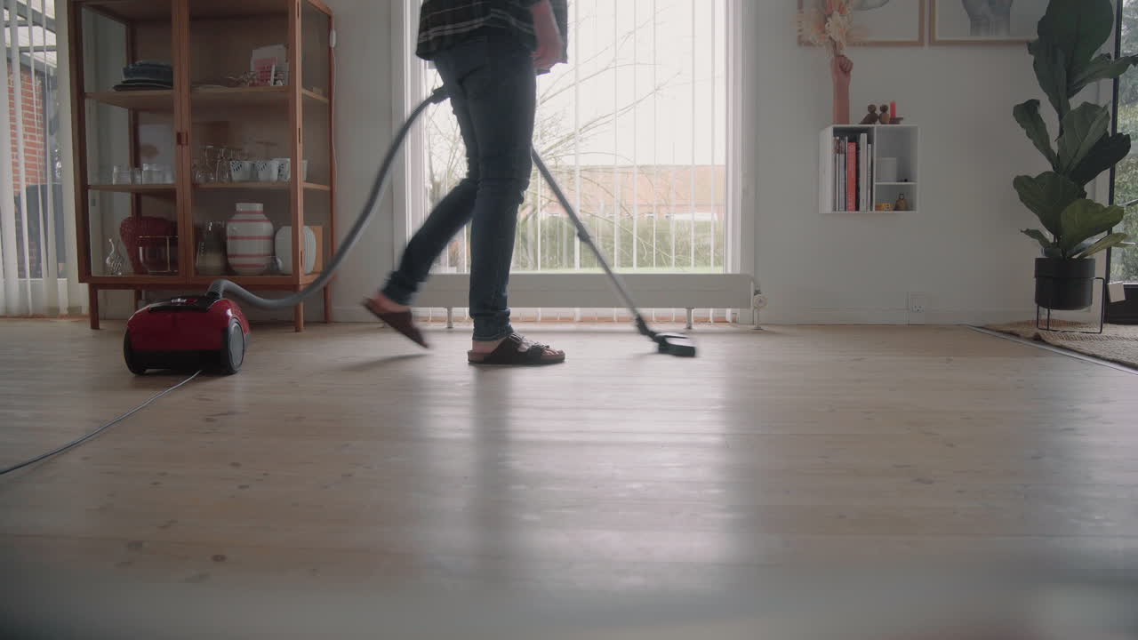 Young hipster male in sandals cleaning the house with a Vacuum Cleaner Cleaning The wooden floor in the living room