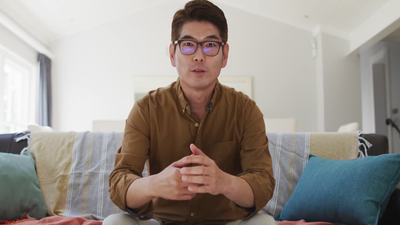Uomo asiatico che lavora da casa, seduto sul divano mentre fa una videochiamata, parla e gesticola.