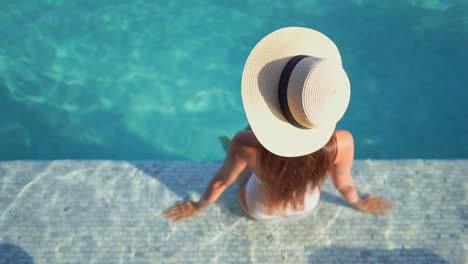 Mujer-En-Traje-De-Baño-Y-Sombrero-De-Verano-Sentada-En-El-Borde-De-Una-Piscina,-Vista-De-ángulo-Alto