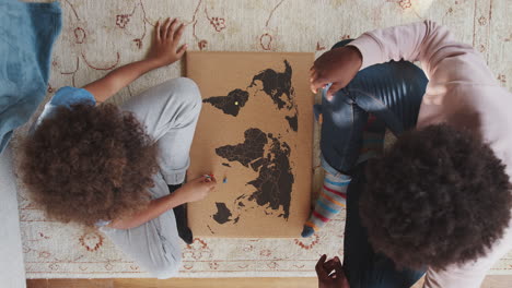 close up overhead view of a mixed race pre teen boy and his father sitting cross legged on the floor opposite each other playing an educational game with a world map and map pins