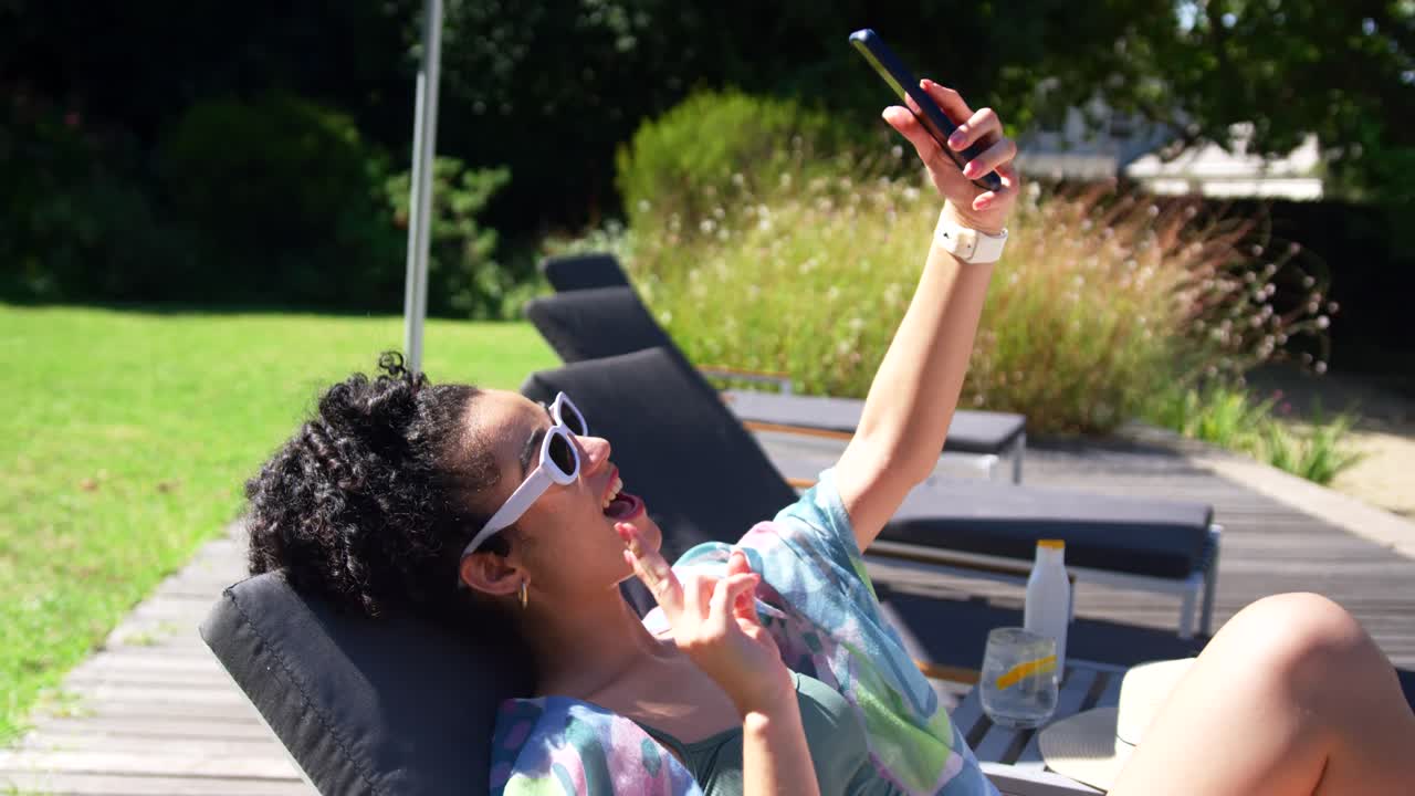 Woman reclining on padded lounge chair on deck, wearing sunglasses, tapping smartphone, talking