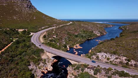 Autos-Conduciendo-Sobre-Un-Fuerte-Puente-De-Concreto-Que-Cruza-Un-Hermoso-Río-Mientras-La-Gente-Nada-Y-Salta-Al-Agua-Con-Montañas-Alrededor-En-Palmiet,-Sudáfrica