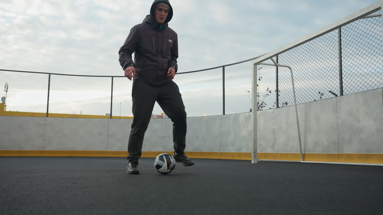 Athlete practicing soccer drills on sport arena, demonstrating footwork skills while controlling ball with precision, background features lamp pole, goalpost, and urban residential buildings