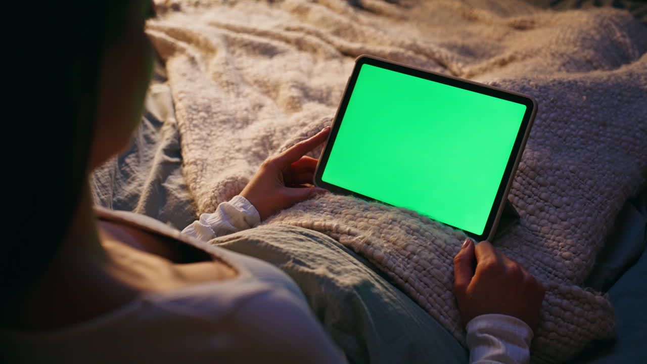 Donna che guarda un tablet con chroma key sdraiata da sola su un letto comodo a casa, primo piano.