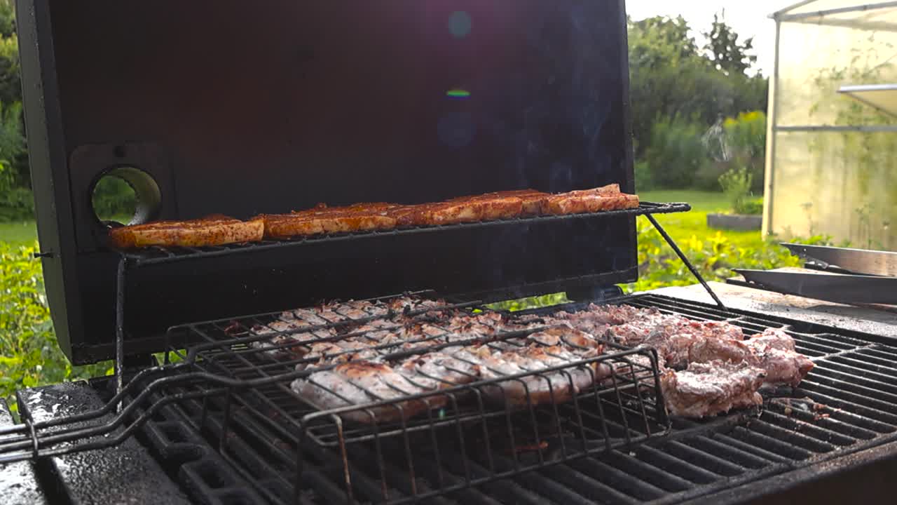 Mans hand with tongs flipping and turning over raw red and brown meat and chicken on a smoking hot grill during a summer sunny day. The meat has char marks and seasoning on it, lense flare in scene