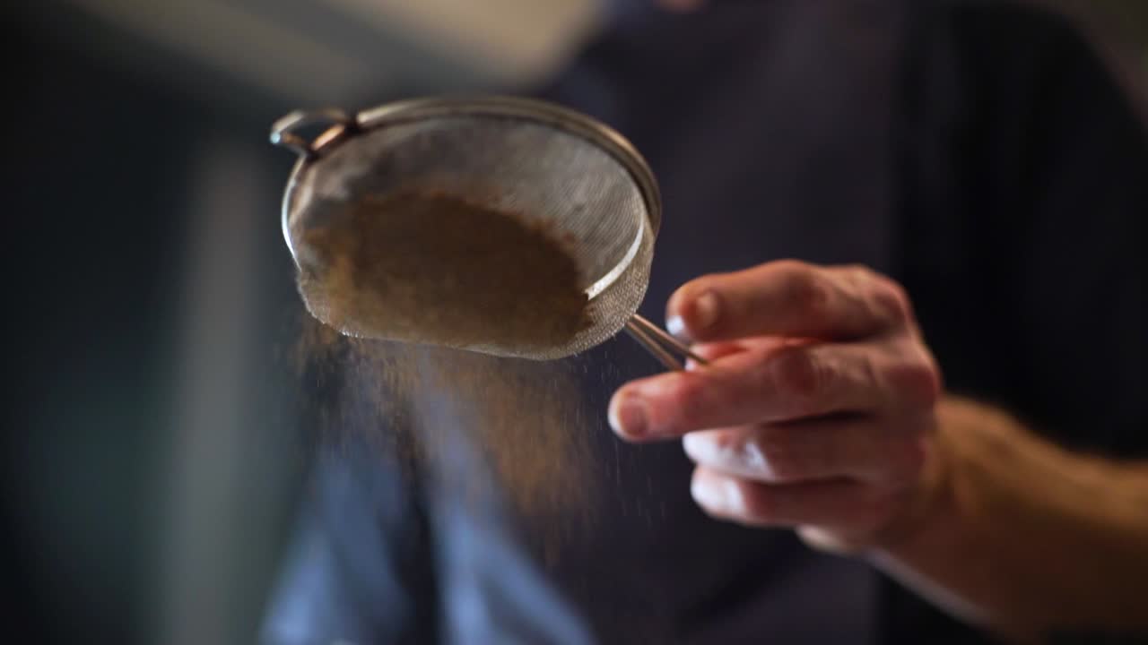 Close up slow motion clip of a chef's hand tapping the handle of a sieve to sprinkle cocoa powder