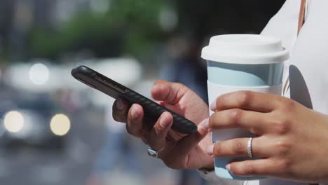 midsection of african american woman using smartphone and holding coffee in street