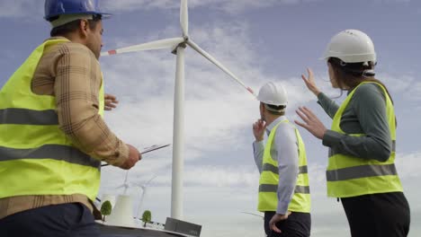 Three-caucasian-and-latin-engineers-standing-on-wind-turbine-field-and-discussing-over-plastic-models.