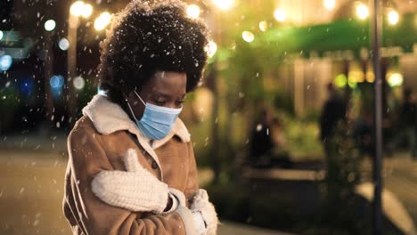 vista de cerca de una mujer afroamericana con máscara facial que tiene frío y trata de calentarse en la calle mientras nieva en navidad