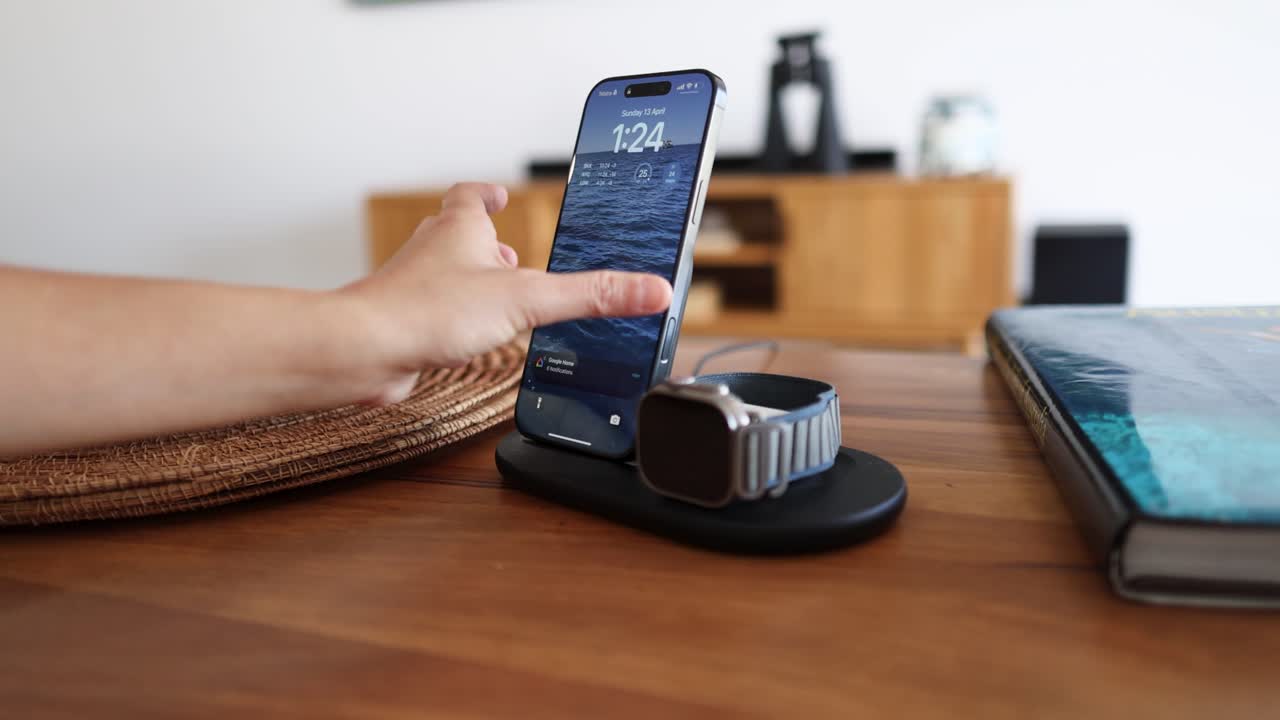 A person places a smartphone on a charging dock beside a smartwatch in a modern home setting with natural lighting
