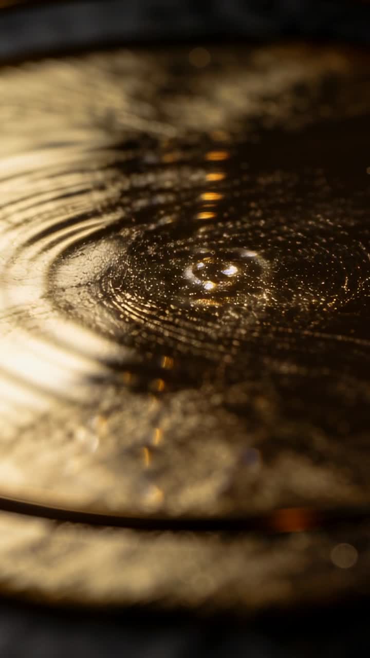 Video verticale: Gocciolina che colpisce un liquido dorato in una ciotola, formando increspature che guidano i glitter per un design.