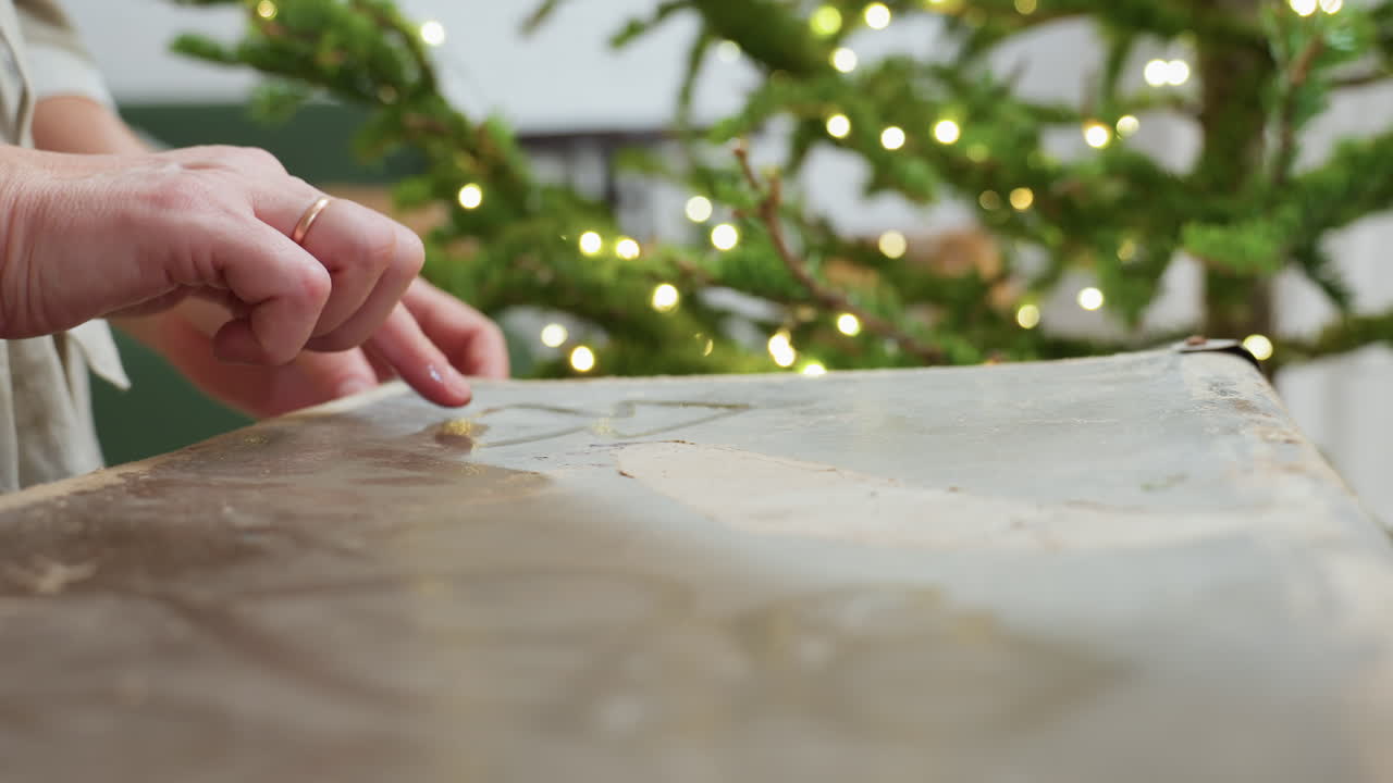 Close hand view of woman drawing playful lines on dusty box surface with finger, creating creative tactile moment during cozy festive indoor atmosphere with blurred Christmas tree in background