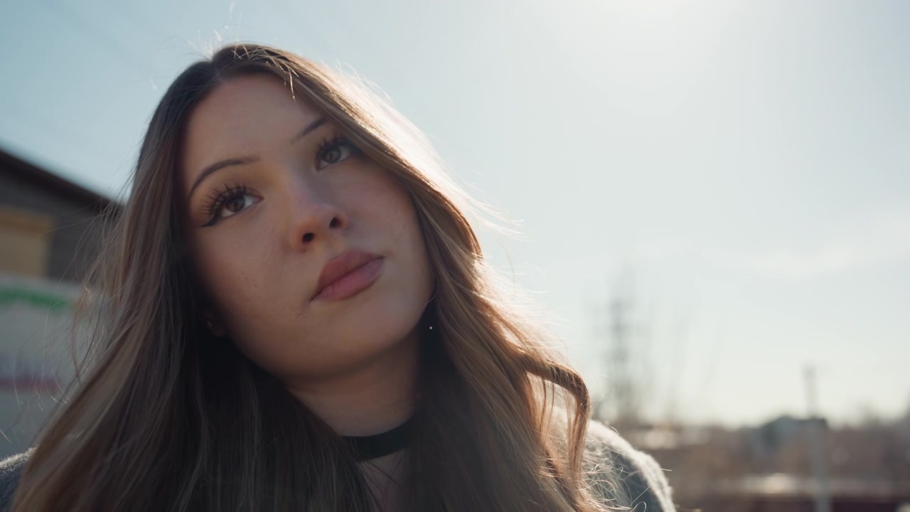 Ritratto all'aperto di una bella donna alla luce del sole, pendolare contemplativa in piedi vicino a un parcheggio urbano, morbida retroilluminazione che evidenzia i capelli lunghi e il cappotto di lana, profondità di campo cinematografica, espressione sottile