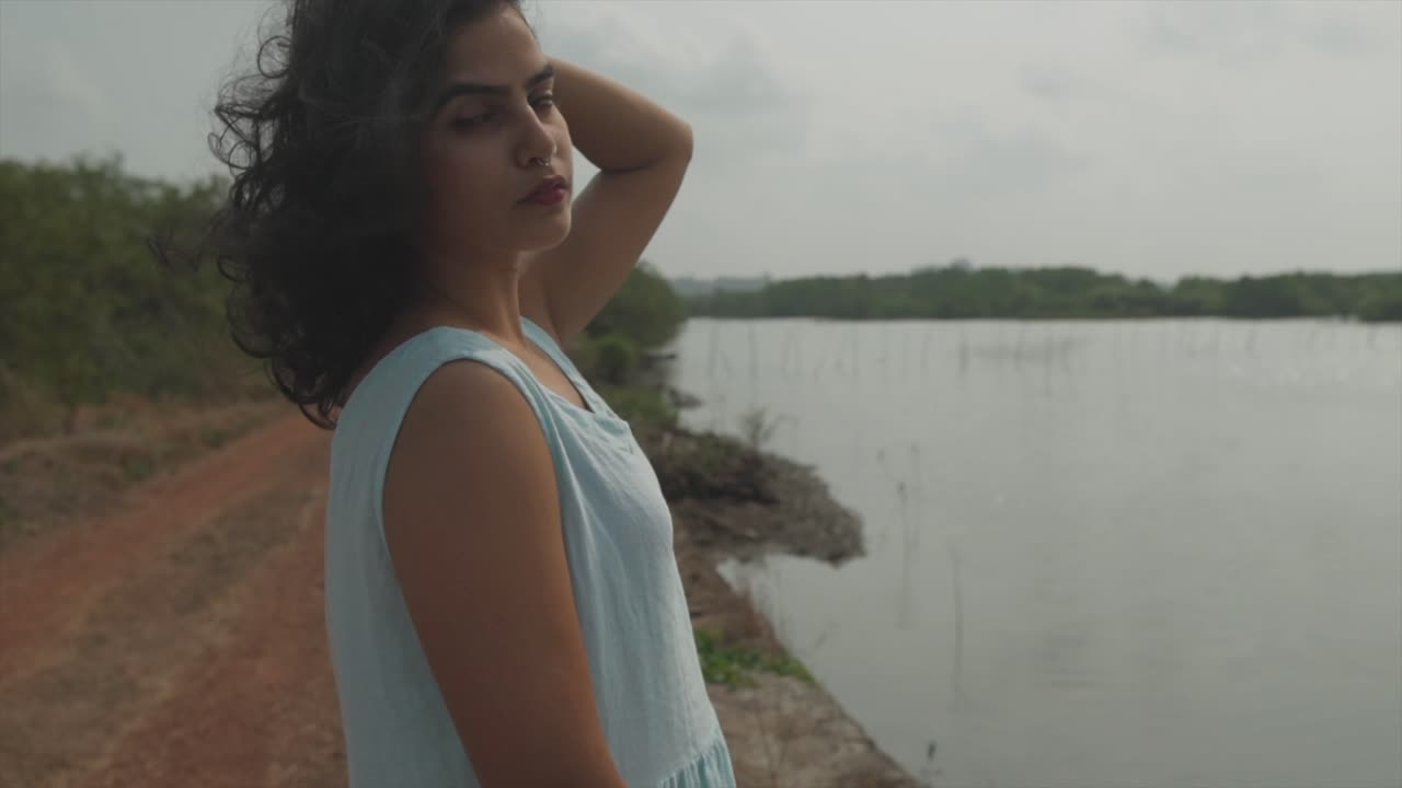 plano medio de una hermosa mujer india vestida con un top azul bebé de pie frente a un lago y mirando a la cámara con una mirada atractiva
