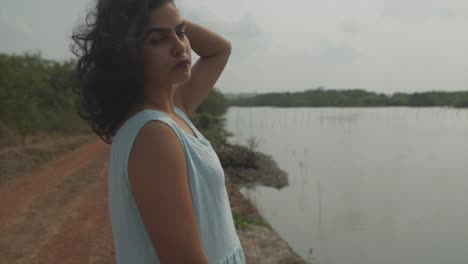 medium shot of a beautiful indian women dressed in a baby blue top standing in front of a lake and looking into the camera with an appealing look