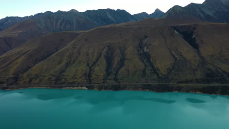 Vista-Aérea-De-Drones-Rastreando-Un-Automóvil-Conduciendo-Por-Una-Pintoresca-Carretera-De-Montaña-A-Lo-Largo-De-Un-Lago-Glacial-Turquesa