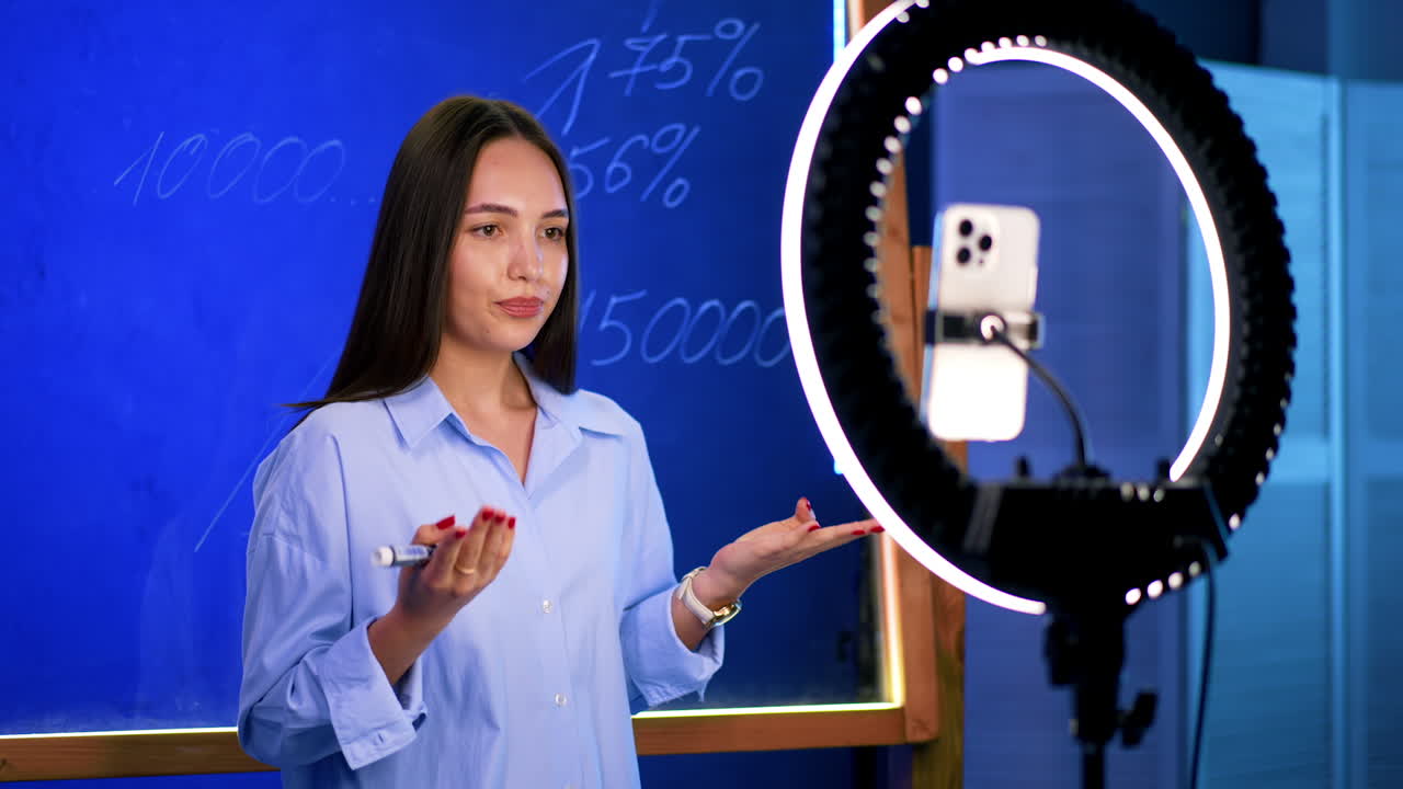 Creative online teaching by youth. A young woman explains a topic while standing in front of a blue background, filming with a smartphone and ring light