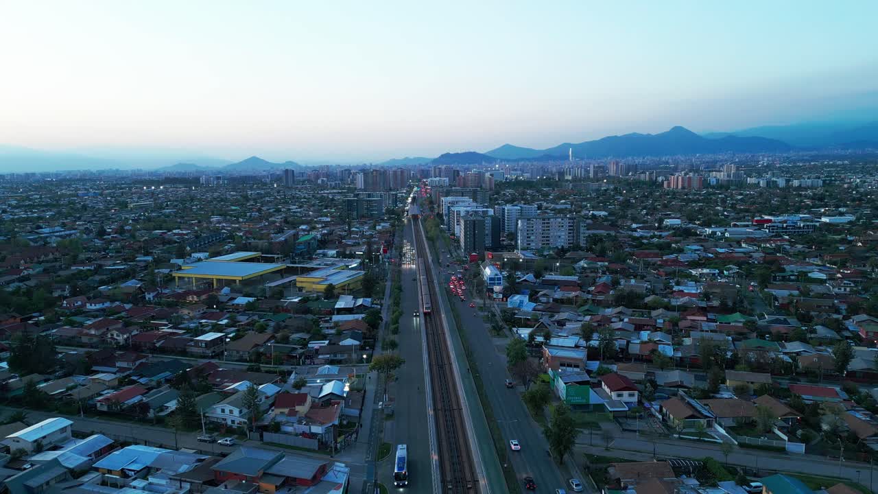 Volo aereo sopra binari ferroviari e autostrada che attraversano un quartiere residenziale con condomini, verso le Ande al tramonto. La Florida, Santiago, Cile.