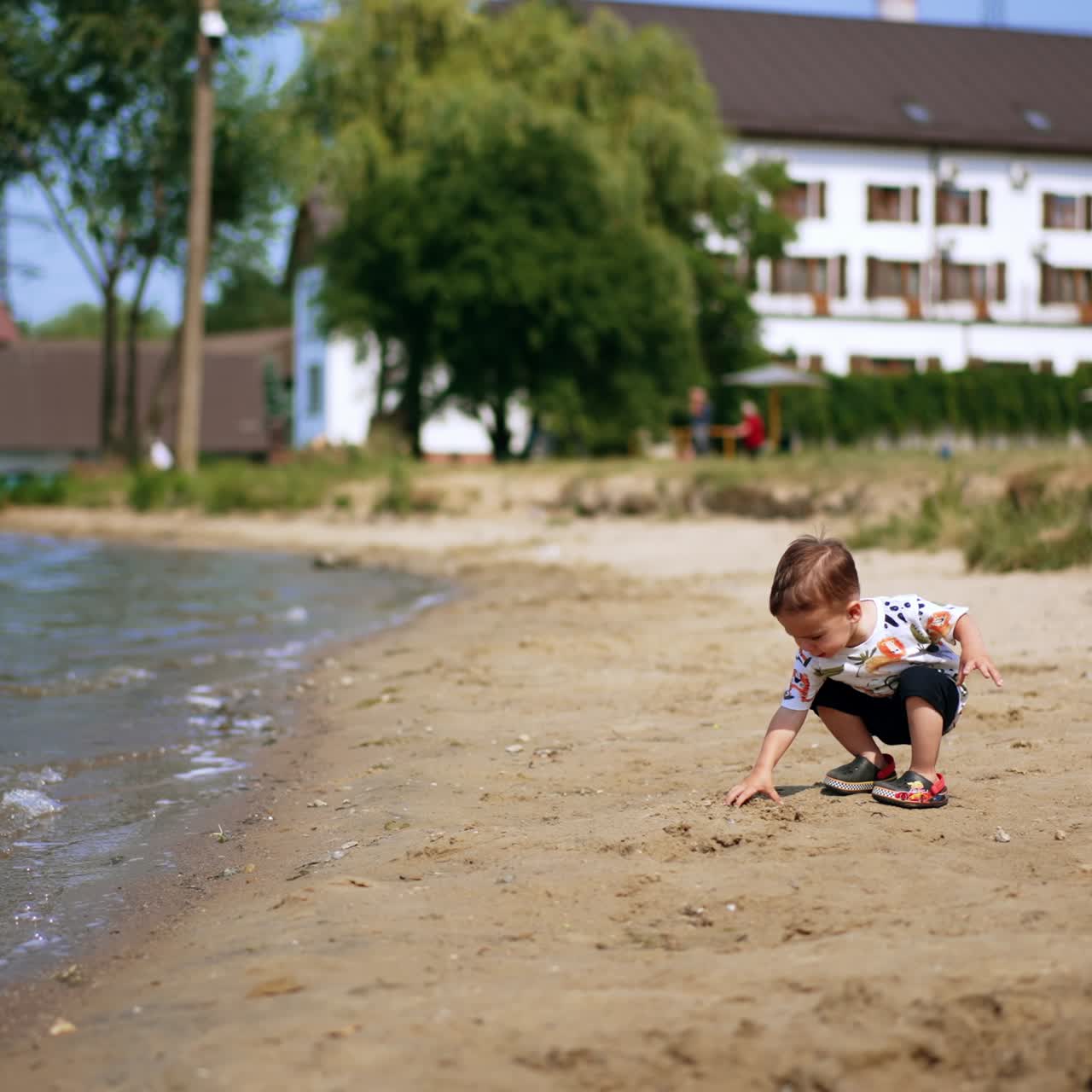 Lovely Caucasian baby standing on the shore. Little boy takes some sand to play with. Summer time joy