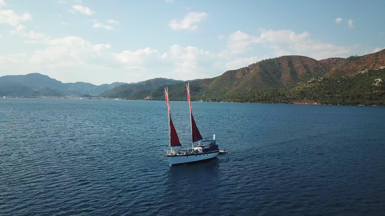 Drone orbit shot of a moving sailing boat with picturesque view of mountains behind.