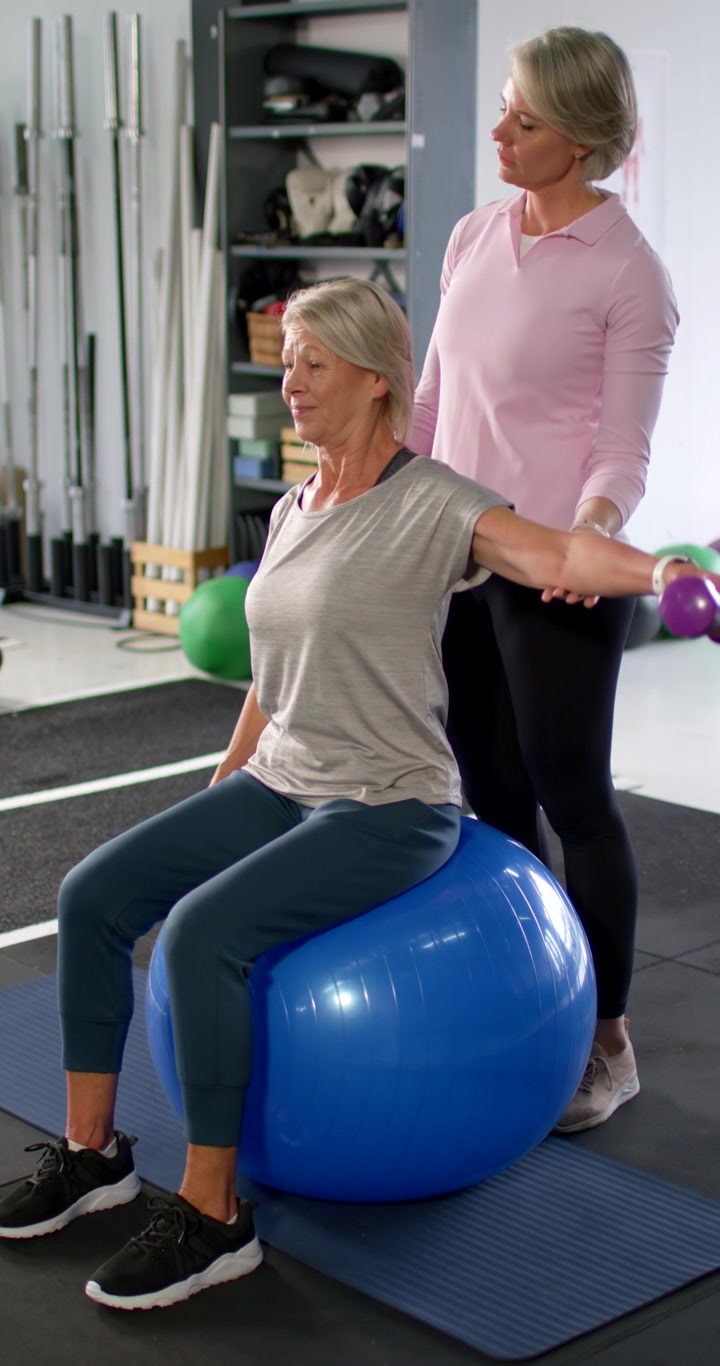 Video verticale: Cliente anziana su una palla blu, con l'allenatore che la stabilizza mentre solleva pesi viola per l'equilibrio.