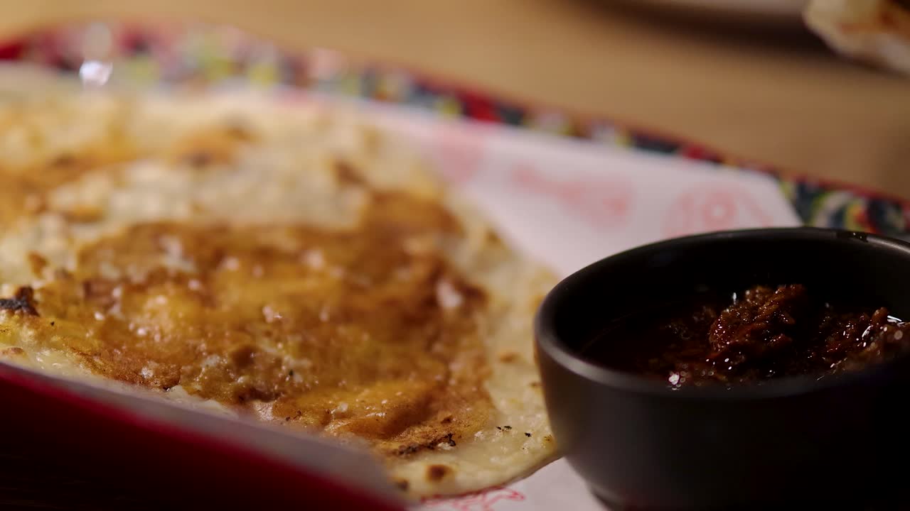 Close-up of a hand tearing grilled flour tortilla and dipping it into spicy sauce under warm indoor lighting, shot with shallow depth of field