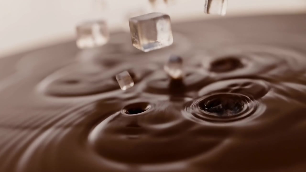 Cubetti di ghiaccio trasparenti cadono in un liquido marrone liscio, creando increspature concentriche su una superficie lucida con tonalità calde beige e cioccolato, riflessi tenui, bassa profondità di campo e uno stile di bevanda premium.
