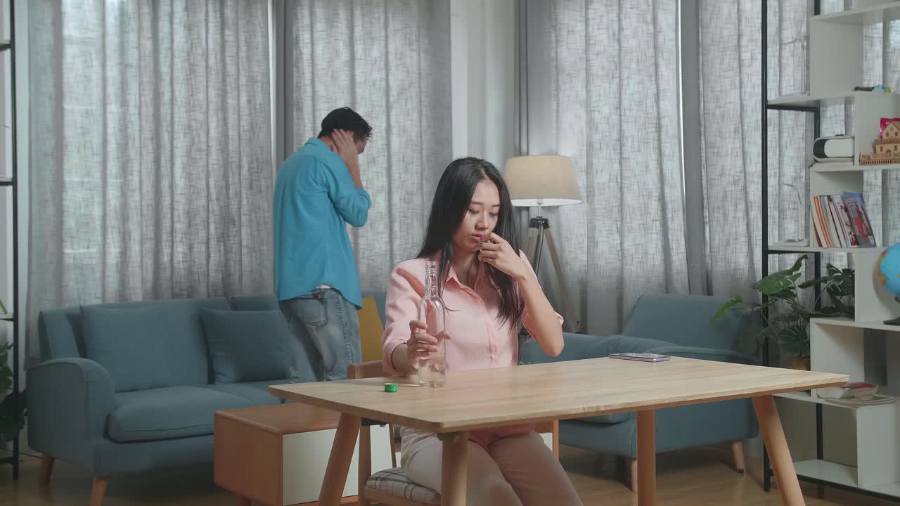 술에 취한 우울한 아시아 여성이 술을 마시기 전에 글라스에 보드카를 부어 울고, 집에서 그녀의 뒤를 따라 는 남자