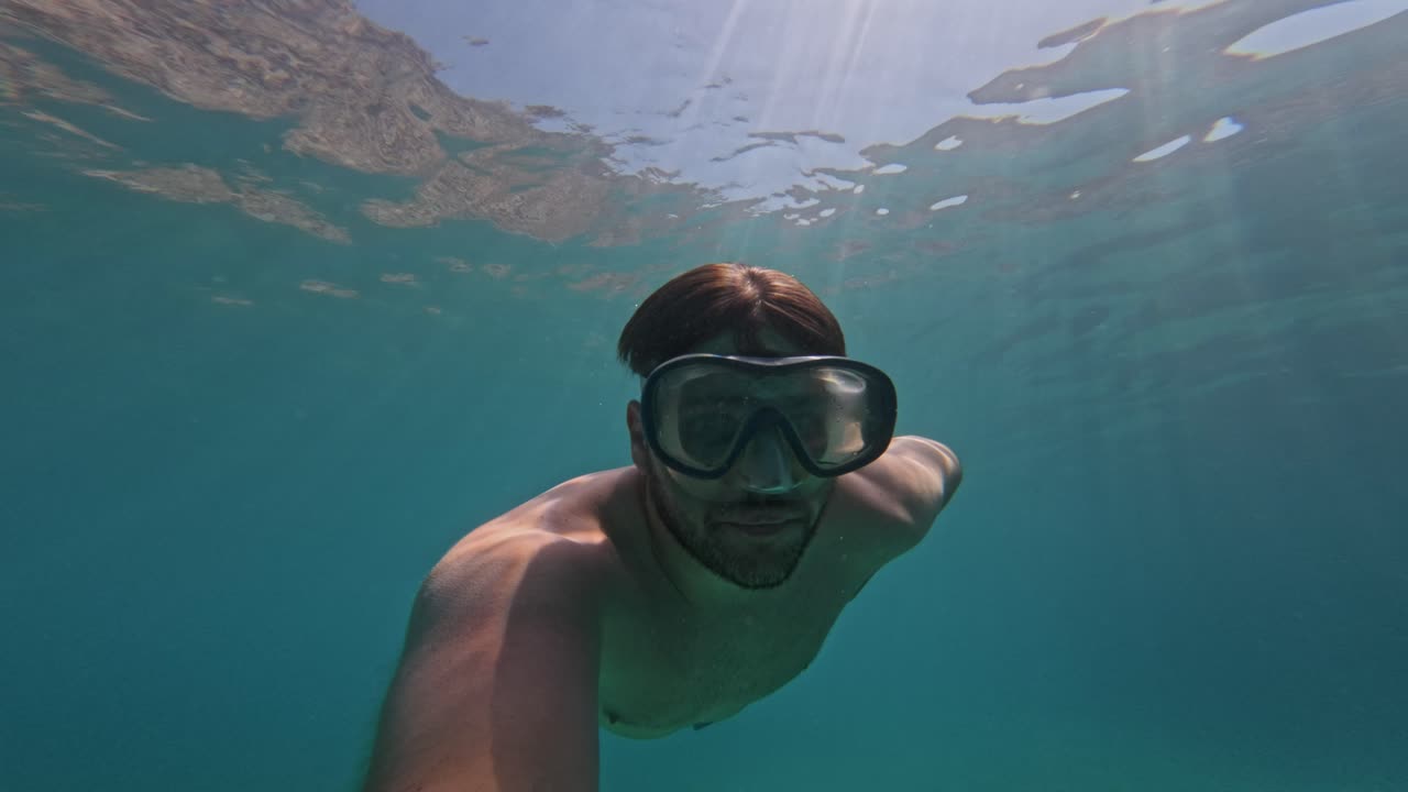 Young Man Wearing Goggles Swimming Under The Sea On A Sunny Day. - underwater shot