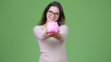 young beautiful woman holding piggy bank against green background