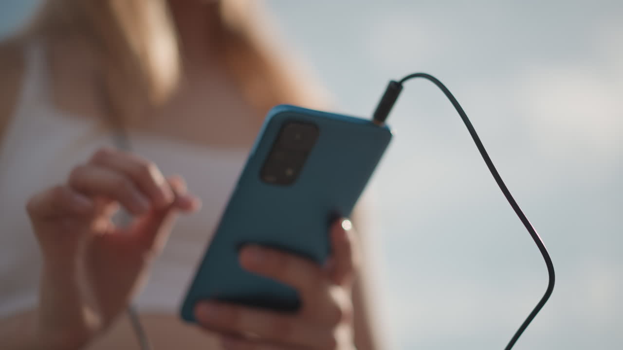 manos sosteniendo un teléfono, bailarina caucásica en exteriores guardando el dispositivo en sus pantalones cortos, ajustando el cable, sintiendo la energía antes de la rutina urbana, horizonte iluminado por el sol, top deportivo recortado, zancada segura, preparación rítmica