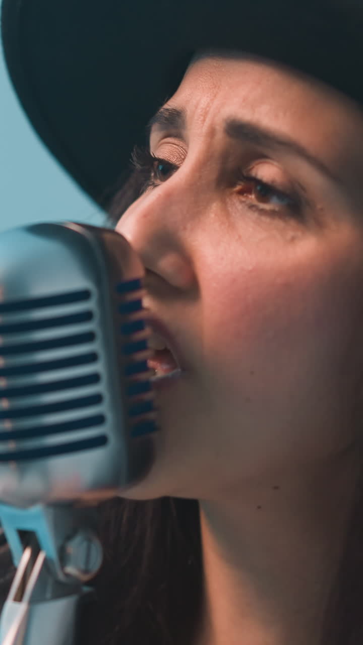 un cantante con un sombrero negro y una chaqueta de cuero negra sostiene un micrófono vintage con una mano mientras gesticula apasionadamente con la otra, en contra de un fondo azul