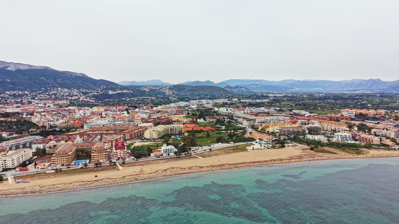 Vista costiera di una città accanto alla spiaggia con montagne sullo sfondo durante tempo nuvoloso.