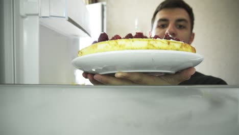 vista desde el refrigerador: el joven abre el refrigerador y pone el pastel decorado con frambuesas