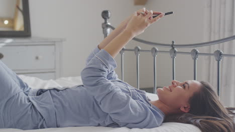 woman wearing pyjamas posing and taking selfies on mobile phone lying on bed