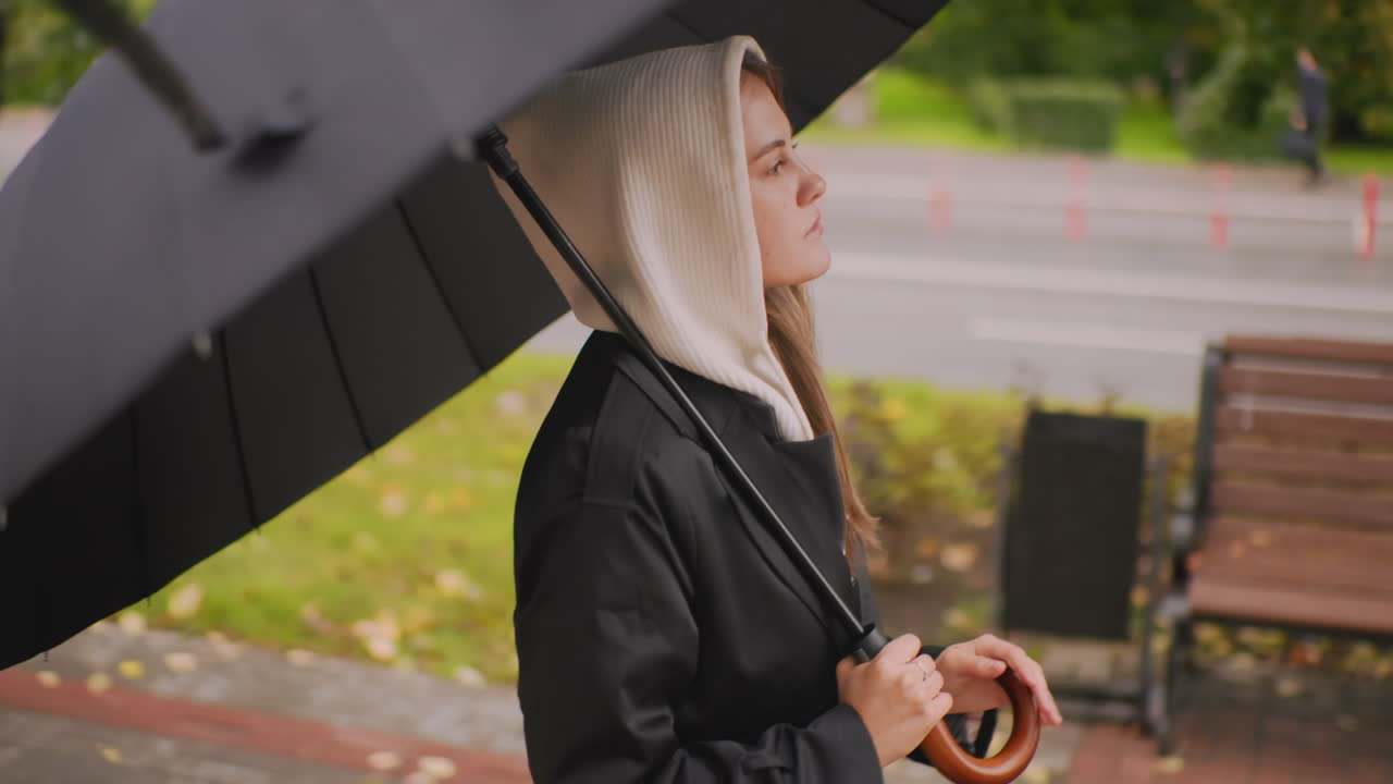 Young woman in hooded coat holding umbrella walking near roadside on rainy day, looking forward with calm expression, autumn leaves on ground, blurred trees and pavement in background, urban lifestyle