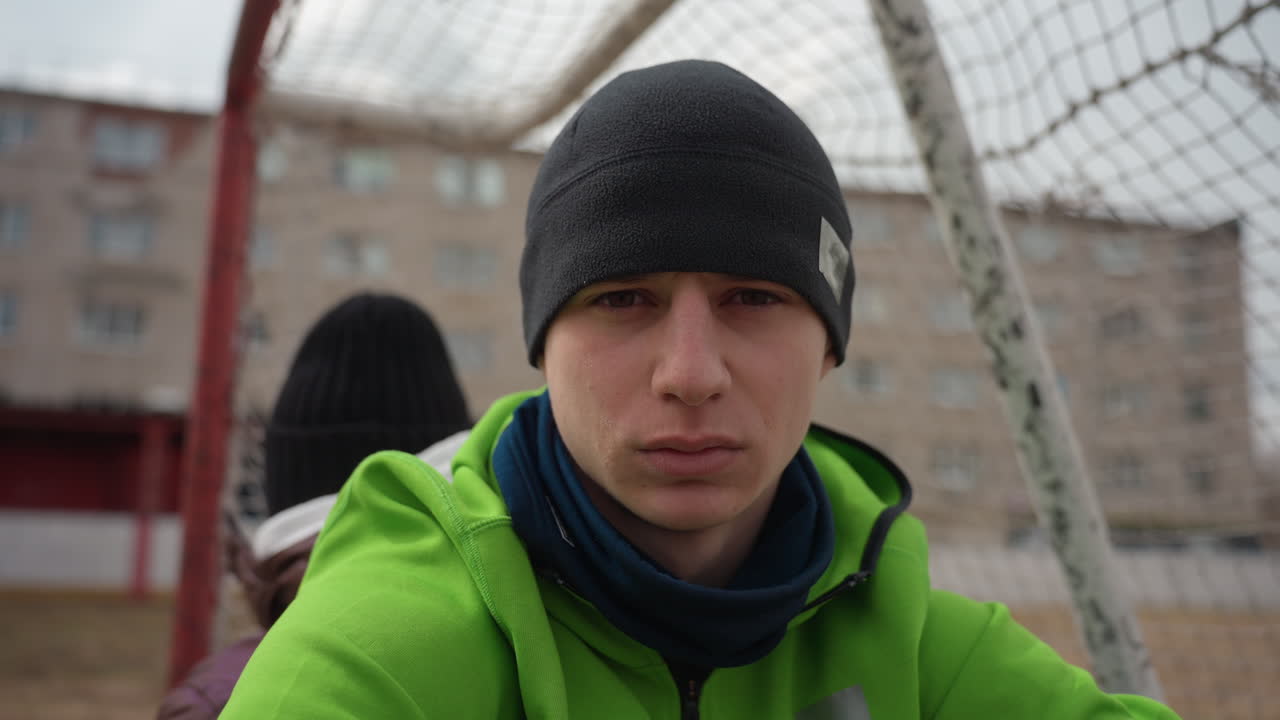 흐린 하늘 아래 도시 경기장에서 골대 그물에 기대어 카메라를 강렬하게 응시하는 백인 골키퍼, 시합 전 준비와 집중을 보여주는 초상화, 초점을 맞춘 시선과 비니