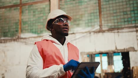 A-man-with-Black-skin-in-a-white-helmet-and-an-orange-vest-stands-at-a-huge-waste-processing-plant-and-writes-data-into-the-tablet-he-is-holding-in-his-hands